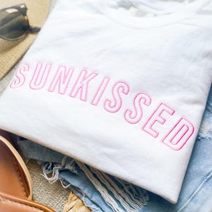May include: A white t-shirt featuring the word SUNKISSED embroidered in pink thread across the chest. The shirt is paired with light blue distressed denim shorts, a straw hat, and brown sandals with the text SHADE SHORE visible on the sole.