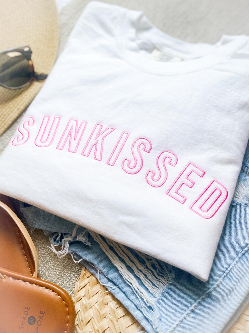 May include: A white t-shirt featuring the word SUNKISSED embroidered in pink thread across the chest. The shirt is paired with light blue distressed denim shorts, a straw hat, and brown sandals with the text SHADE SHORE visible on the sole.