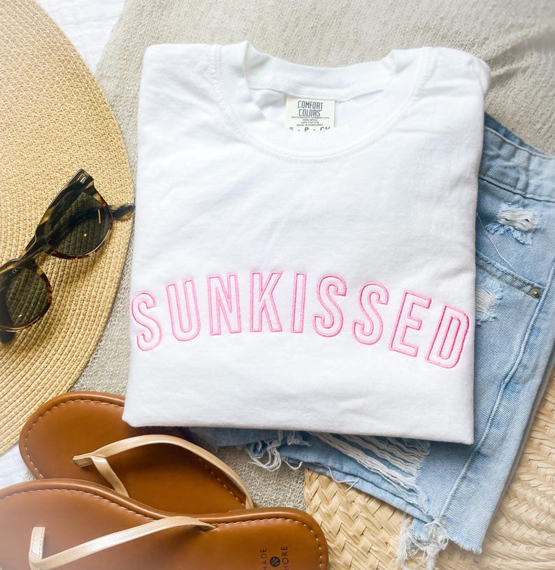 May include: A white short-sleeved t-shirt with the word SUNKISSED embroidered in pink capital letters across the chest. The shirt features a crew neckline and a Comfort Colors brand label on the inside of the collar.