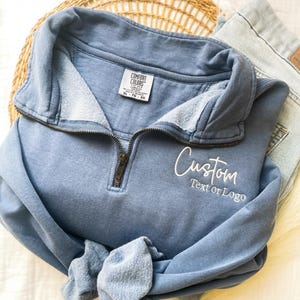 May include: A blue quarter-zip sweatshirt featuring a Comfort Colors brand label inside the collar and white embroidered text on the chest that reads Custom Text or Logo. The garment has a metal zipper pull and a soft, fleece-lined interior.