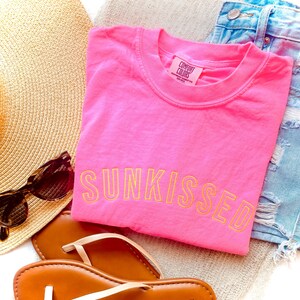 May include: A bright pink short-sleeved t-shirt with the word SUNKISSED embroidered in yellow thread across the chest. The shirt features a Comfort Colors label at the inner neckline. The garment is displayed alongside a straw hat and sandals.