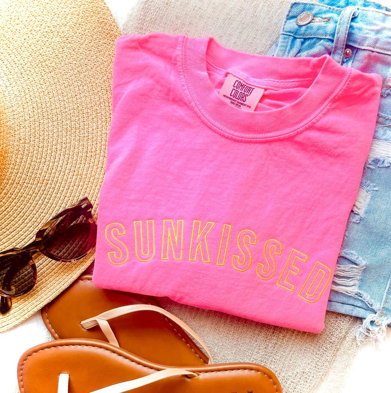 May include: A bright pink short-sleeved t-shirt with the word SUNKISSED embroidered in yellow thread across the chest. The shirt features a Comfort Colors label at the inner neckline. The garment is displayed alongside a straw hat and sandals.