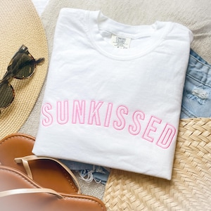 May include: A white short-sleeved t-shirt featuring the word SUNKISSED embroidered in pink capital letters across the chest. The shirt has a crew neckline and a Comfort Colors brand label visible at the inner collar area.