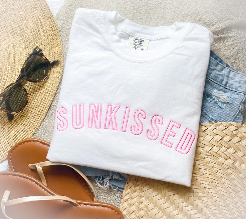 May include: A white short-sleeved t-shirt featuring the word SUNKISSED embroidered in pink capital letters across the chest. The shirt has a crew neckline and a Comfort Colors brand label visible at the inner collar area.