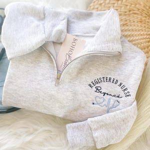 May include: A light gray, quarter-zip sweatshirt with the words "REGISTERED NURSE Beyonce" embroidered in black and light blue. A stethoscope graphic is also included. A tag that says "Boho Barn" is visible. A stethoscope and denim are in the background.