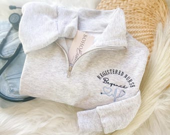 May include: A light gray, quarter-zip sweatshirt with the words "REGISTERED NURSE Beyonce" embroidered in black and light blue. A stethoscope graphic is also included. A tag that says "Boho Barn" is visible. A stethoscope and denim are in the background.