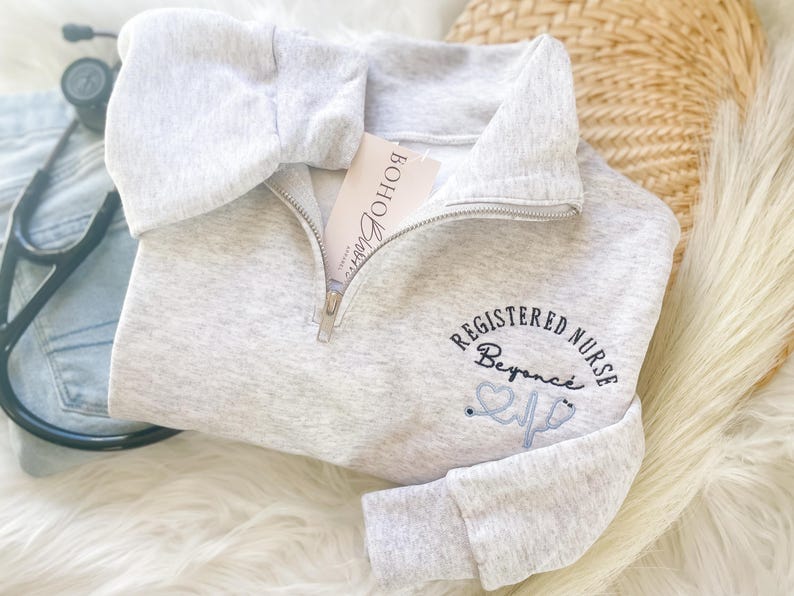 May include: A light gray, quarter-zip sweatshirt with the words "REGISTERED NURSE Beyonce" embroidered in black and light blue. A stethoscope graphic is also included. A tag that says "Boho Barn" is visible. A stethoscope and denim are in the background.