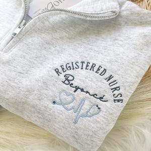 May include: A light gray quarter-zip sweatshirt with the words "REGISTERED NURSE" embroidered in navy blue, along with the name "Beyonc&eacute;" and a light blue stethoscope design. A white tag with "BOHO APPAREL" is attached.