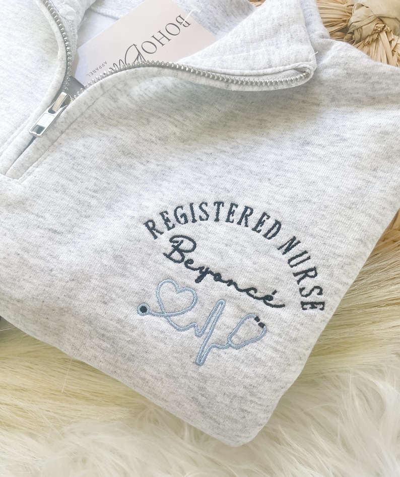May include: A light gray quarter-zip sweatshirt with the words "REGISTERED NURSE" embroidered in navy blue, along with the name "Beyonc&eacute;" and a light blue stethoscope design. A white tag with "BOHO APPAREL" is attached.