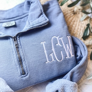 May include: A blue quarter-zip sweatshirt featuring a white embroidered monogram with the letters LCW on the left chest. The garment has a metal zipper closure, a stand-up collar, and a visible interior tag indicating size M.