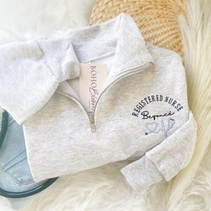 May include: A light gray quarter-zip sweatshirt with the text "REGISTERED NURSE Beyonce" embroidered in black and light blue. A stethoscope and a tag that says "Boho Blossoms" are also visible.