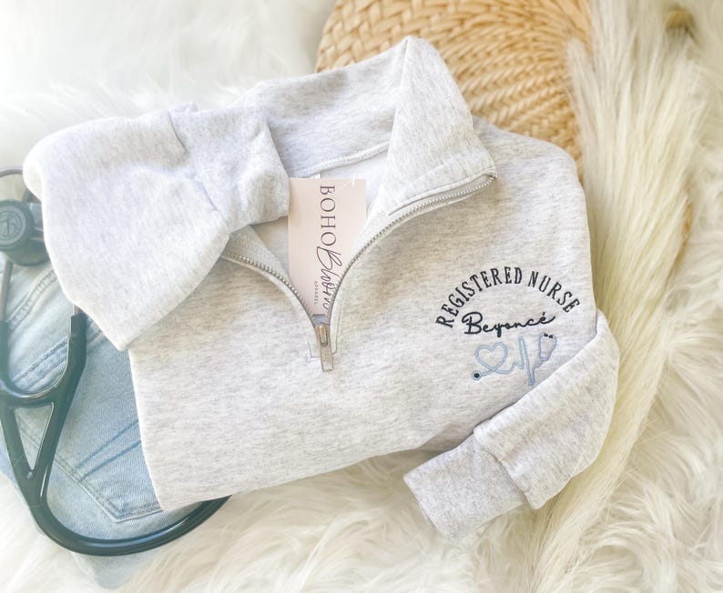 May include: A light gray quarter-zip sweatshirt with the text "REGISTERED NURSE Beyonce" embroidered in black and light blue. A stethoscope and a tag that says "Boho Blossoms" are also visible.
