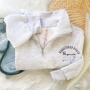 May include: A light gray quarter-zip sweatshirt with the text "REGISTERED NURSE Beyonce" embroidered in black and light blue. A stethoscope and a tag that says "Boho Bloom" are also visible.