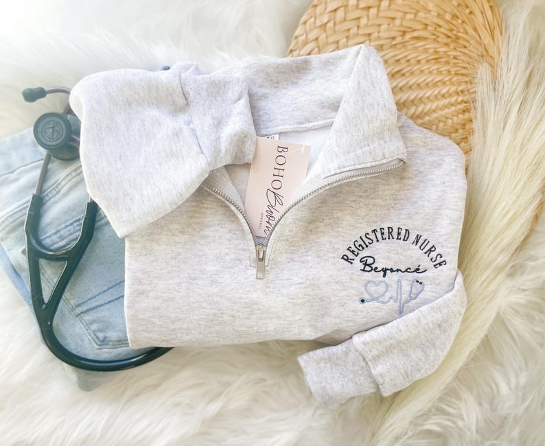 May include: A light gray quarter-zip sweatshirt with the text "REGISTERED NURSE Beyonce" embroidered in black and light blue. A stethoscope and a tag that says "Boho Bloom" are also visible.
