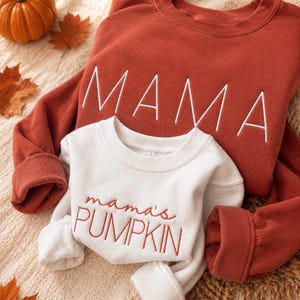 May include: A rust-coloured long-sleeve sweatshirt with the word MAMA embroidered in white thread, layered with a smaller white long-sleeve sweatshirt featuring the embroidered text mama's PUMPKIN in rust-coloured thread on the front.