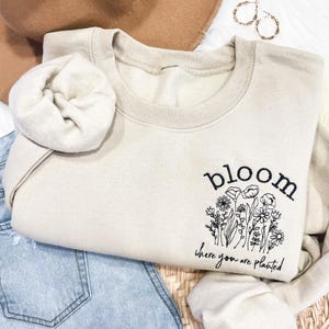 May include: A beige sweatshirt with the text "bloom where you are planted" and a black line drawing of flowers.