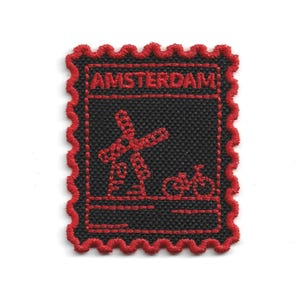 May include: Embroidered patch featuring a black background with a red windmill and bicycle design. The word "AMSTERDAM" is stitched across the top. The patch has a red, scalloped border.