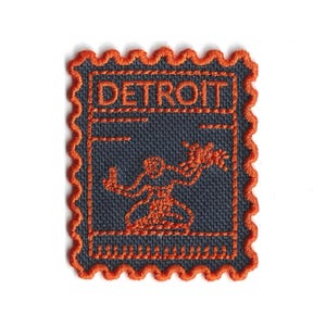 May include: Embroidered patch in a postage stamp design. The patch features the word "DETROIT" in orange thread, along with an orange figure holding a bouquet. The background is a dark blue fabric, and the edges are scalloped in orange.