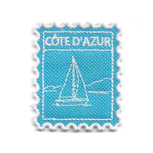 May include: A rectangular embroidered patch with a scalloped white border on a bright blue fabric background. The design features a white stitched sailboat on water and the text COTE D'AZUR embroidered in white at the top of the patch.