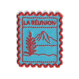 Könnte beinhalten: Gestickter Aufnäher mit dem Text "LA RÉUNION" über einem Berg- und Pflanzendesign. Der Aufnäher hat einen hellblauen Hintergrund mit roten Stickdetails und einem roten, gewellten Rand. Der Aufnäher ist quadratisch.