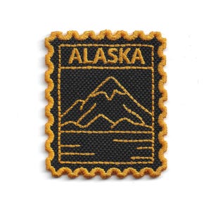 Peut inclure: Un écusson noir avec des coutures dorées portant le mot "ALASKA" en haut. En dessous, un motif de chaîne de montagnes. L'écusson a une bordure dorée festonnée, ressemblant à un timbre-poste.