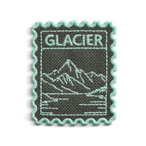 Può includere: Una toppa ricamata rettangolare con bordo smerlato in filo azzurro. Il tessuto grigio scuro presenta un paesaggio montano cucito in azzurro e la parola GLACIER in lettere maiuscole nella parte superiore, simile a un francobollo.
