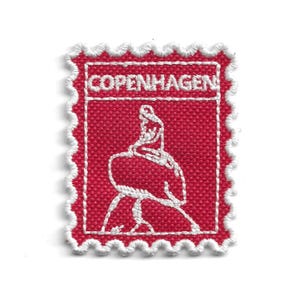 以下が含まれることがあります： 切手型の赤い刺繍パッチ。白いステッチが施され、上部に「COPENHAGEN」の文字があります。中央には彫像の白いアウトラインがあります。パッチには白いスカラップのボーダーが付いています。