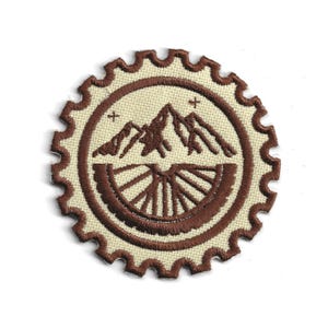 May include: Round, embroidered patch featuring a mountain range and a bicycle wheel design in shades of brown and beige. The patch has a scalloped edge and is likely for a backpack or jacket.