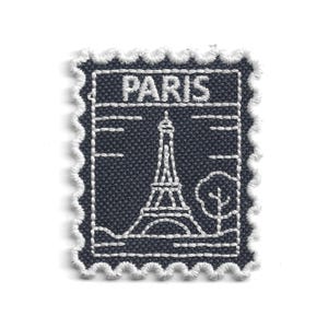 Pode incluir: Um patch de tecido azul marinho em forma de selo postal. O patch apresenta a palavra "PARIS" no topo, com um design bordado branco da Torre Eiffel e uma árvore. As bordas são recortadas com fio branco.