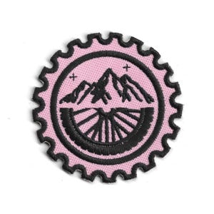 May include: A pink embroidered patch with a black mountain and wheel design. The patch has a scalloped edge and two small crosses. The mountain design is in the center of a black circle.