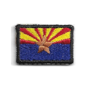 May include: A rectangular embroidered patch featuring the Arizona state flag design. The top half displays alternating red and yellow sun rays, while the bottom half is solid blue. A large, five-pointed copper-colored star is centered.