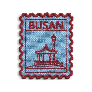 Puede incluir: Un parche bordado rectangular con un borde festoneado rojo y un fondo de tela azul claro. La palabra BUSAN está bordada en rojo en la parte superior. Debajo del texto, un contorno rojo representa un pabellón tradicional y una farola alta.