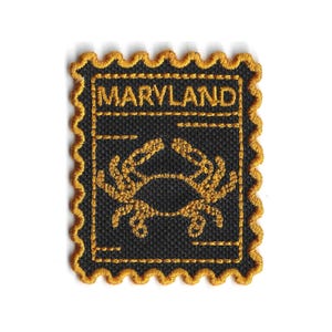 Könnte beinhalten: Gestickter Aufnäher mit schwarzem Hintergrund, goldenem Krabbenmuster und dem Wort "MARYLAND" in Gold. Der Aufnäher hat einen goldfarbenen, gewellten Rand, der einer Briefmarke ähnelt. Ideal zum Aufnähen auf Jacken, Taschen oder andere Stoffartikel.