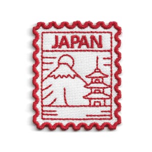 May include: A rectangular embroidered patch with a scalloped red border on a white fabric base. The design features the word JAPAN in red capital letters above a red line-art illustration of a mountain and a multi-tiered pagoda structure.