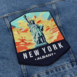 Pode incluir: Um patch retangular com a Estátua da Liberdade contra um céu colorido, costurado em uma jaqueta jeans. O patch tem as palavras "NEW YORK" e "ALBANY".