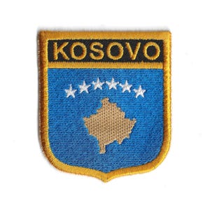 May include: Embroidered patch featuring the word "KOSOVO" in gold lettering above a blue shield. The shield displays five white stars and a tan outline of the country. The patch has a gold border.