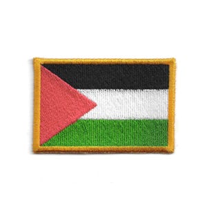 Op de afbeelding: Geborduurde patch met de vlag van Palestina. Het ontwerp toont horizontale strepen in zwart, wit en groen, met een rode driehoek aan de linkerkant. De patch heeft een gouden rand en is rechthoekig.