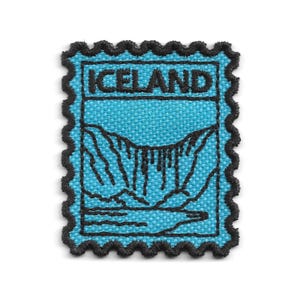 Peut inclure: Un écusson brodé rectangulaire avec une bordure noire festonnée, présentant un fond bleu clair et des détails en fil noir. Le motif représente un paysage de cascade avec le mot ICELAND brodé en noir sur la partie supérieure de l'écusson.