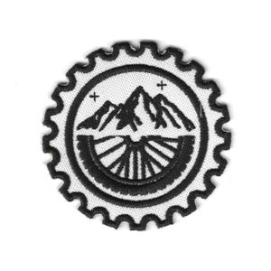 May include: A circular black and white embroidered patch featuring a mountain range and a stylized sun or wheel design. The patch has a scalloped edge and two small cross symbols. The design is centered on a white background.