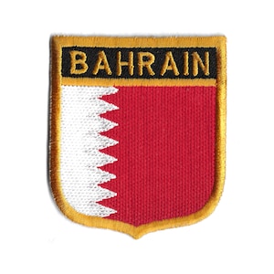 Puede incluir: Un parche bordado rojo y blanco con la palabra "BAHRAIN" en letras doradas sobre fondo negro. El parche tiene forma de escudo con un borde dorado.