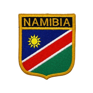 May include: Embroidered patch featuring the word "NAMIBIA" in gold lettering above a shield design. The shield displays a blue field with a yellow sun, a red diagonal stripe, and a green lower section, all bordered in gold.