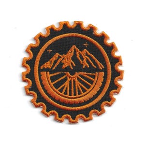 May include: Embroidered patch featuring a mountain range and a bicycle wheel design in orange and black. The patch has a circular shape with a gear-like border. Two small stars are above the mountain range.