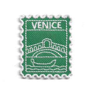 Puede incluir: Un parche de tela verde con un diseño bordado en blanco de un puente y una góndola, con la palabra "VENICE" en la parte superior. El parche tiene un borde festoneado.