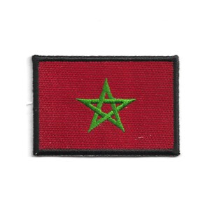 Puede incluir: Un parche rectangular con la bandera de Marruecos. El parche tiene un fondo rojo con una estrella verde de cinco puntas en el centro. El borde es negro. Este parche es probablemente para ropa o accesorios.