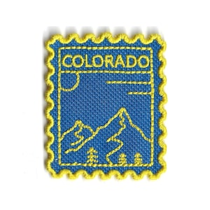 Puede incluir: Parche bordado con diseño de sello postal con la palabra "COLORADO" en la parte superior. El parche tiene un fondo azul con costuras amarillas, que representa una cordillera, árboles y un sol. El parche tiene un borde amarillo festoneado.
