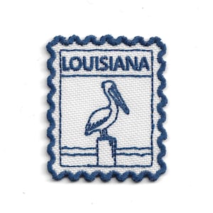 May include: A rectangular embroidered patch with a scalloped edge, featuring a white background and dark blue stitching. The design depicts a pelican perched on a post above water, with the word LOUISIANA embroidered in dark blue at the top.