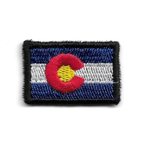 Può includere: Toppa ricamata con la bandiera dello stato del Colorado. La toppa rettangolare ha uno sfondo blu con una striscia bianca e una 'C' rossa con un centro giallo. La toppa è bordata con cuciture nere.