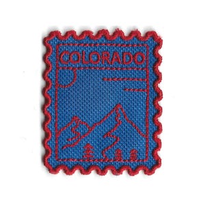 Op de afbeelding: Een blauwe patch met een rode rand en het woord "COLORADO" geborduurd. De patch heeft een bergketen, bomen en een zonontwerp. De patch is rechthoekig met een geschulpte rand.