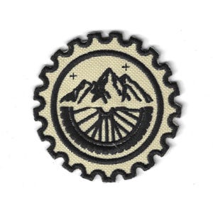 Könnte beinhalten: Ein kreisförmiger Aufnäher mit einem Bergketten- und Fahrradfelgen-Design. Der Aufnäher hat einen cremefarbenen Hintergrund mit einem schwarzen Rand und einer schwarzen Berg- und Felgengrafik. Zwei kleine Pluszeichen befinden sich über den Bergen.