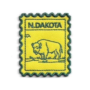 May include: A yellow embroidered patch with a green scalloped border. The patch features the text "N.DAKOTA" at the top and a green outline of a bison. The background is a textured yellow fabric.